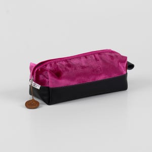 Puede incluir: Un estuche para lápices de terciopelo rosa y cuero negro con cierre de cremallera y una etiqueta de cuero marrón que dice "Something Special".