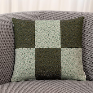 Op de afbeelding: Vierkant decoratief kussen met een groen en lichtgroen geruit patroon. Het kussen is gemaakt van een textuurstof en ligt op een grijze bank. Er is een klein label zichtbaar aan de zijkant.