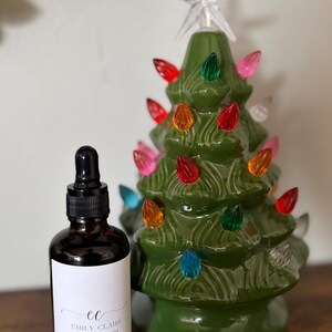 May include: A green ceramic Christmas tree with colorful lights and a star topper. Next to it is a dark glass bottle with a dropper and a white label that reads "Emily Claire Bath and Body."