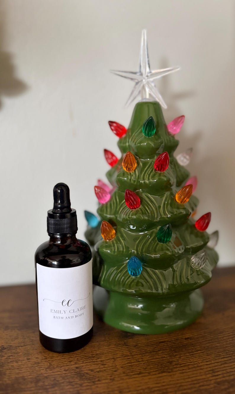 May include: A green ceramic Christmas tree with colorful lights and a star topper. Next to it is a dark glass bottle with a dropper and a white label that reads "Emily Claire Bath and Body."