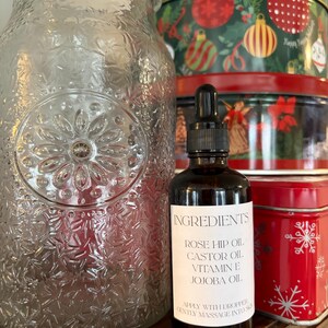 May include: A dark glass bottle with a dropper, labeled "INGREDIENTS" with a list of oils. A large, clear glass jar with a floral design is to the left. Christmas tins are in the background.