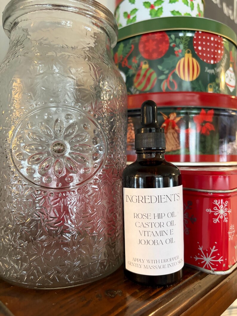 May include: A dark glass bottle with a dropper, labeled "INGREDIENTS" with a list of oils. A large, clear glass jar with a floral design is to the left. Christmas tins are in the background.