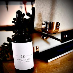 May include: A dark brown glass bottle with a dropper, featuring a white label with the text "ec EMILY CLAIRE BATH AND BODY". The bottle is set against a backdrop of a white textured surface and greenery.