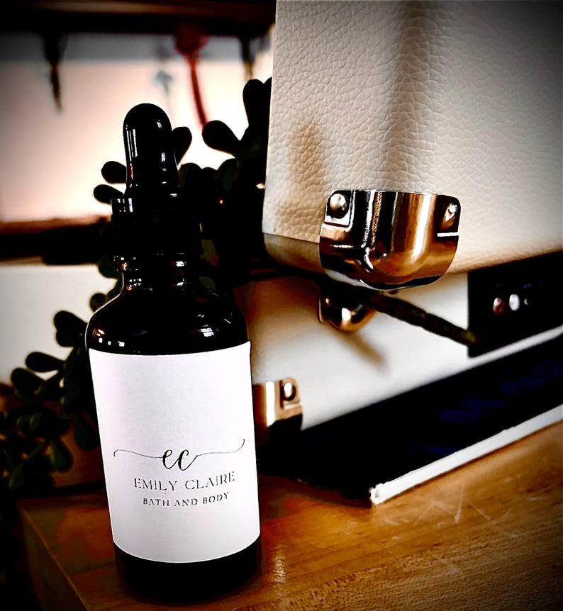 May include: A dark brown glass bottle with a dropper, featuring a white label with the text "ec EMILY CLAIRE BATH AND BODY". The bottle is set against a backdrop of a white textured surface and greenery.