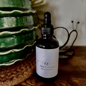 May include: A dark brown glass bottle with a black dropper and a white label. The label reads "ec EMILY CLAIRE BATH AND BODY" in a script font. The bottle is on a wooden surface, with a green ceramic vase and a metal snail decoration in the background.