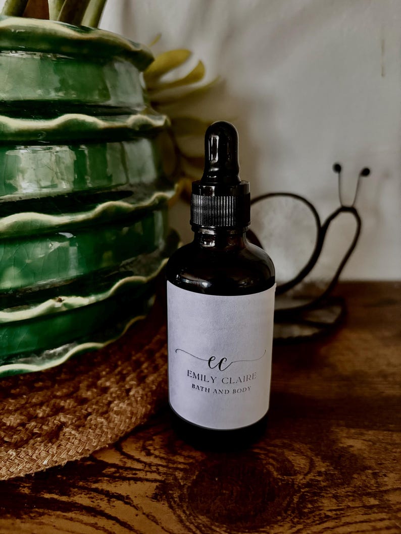 May include: A dark brown glass bottle with a black dropper and a white label. The label reads "ec EMILY CLAIRE BATH AND BODY" in a script font. The bottle is on a wooden surface, with a green ceramic vase and a metal snail decoration in the background.