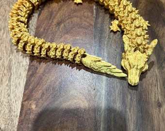 May include: A golden-yellow, articulated dragon figurine. The dragon is made of many small, textured segments, with a detailed head and tail. The dragon is posed in a curved shape, resting on a wooden surface.