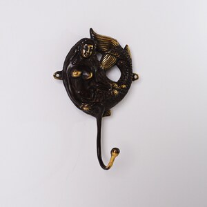 May include: A dark bronze-toned metal wall hook featuring a sculpted mermaid figure with a curved tail that forms a circular frame. The mermaid has polished gold-coloured highlights on her hair, shoulders, and tail, with a single hook extending below.