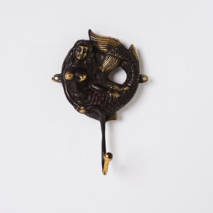 May include: A dark bronze-toned metal wall hook featuring a mermaid figure with a curled tail. The mermaid has polished gold-toned highlights on her hair, chest, and tail fin. The hook extends downwards from the base of the mermaid's tail.