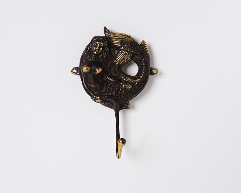 May include: A dark bronze-toned metal wall hook featuring a mermaid figure with a curled tail. The mermaid has polished gold-toned highlights on her hair, chest, and tail fin. The hook extends downwards from the base of the mermaid's tail.