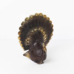 May include: A brass peacock figurine featuring a dark, textured body and a fanned tail with polished gold-toned circular accents. The metal sculpture displays intricate feather detailing across the plumage and a smooth, pointed beak on the head.