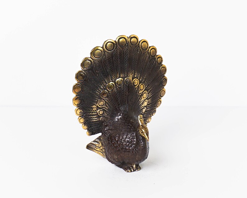 May include: A brass peacock figurine featuring a dark, textured body and a fanned tail with polished gold-toned circular accents. The metal sculpture displays intricate feather detailing across the plumage and a smooth, pointed beak on the head.