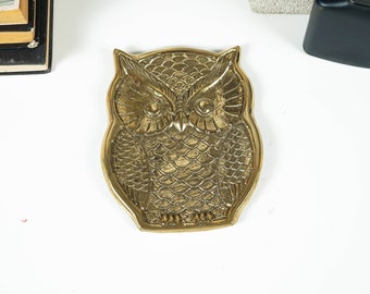 May include: A brass owl-shaped tray featuring a detailed embossed feather pattern across the body and wings. The metal dish has a polished gold-tone finish with defined facial features, including large circular eyes and a pointed beak.