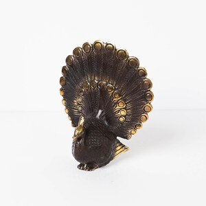 May include: A metallic peacock figurine featuring a dark bronze-toned body and a fanned tail with raised, polished gold-colored circular accents. The bird is depicted in a standing position with its head turned slightly to the side and wings tucked.