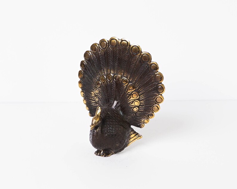 May include: A metallic peacock figurine featuring a dark bronze-toned body and a fanned tail with raised, polished gold-colored circular accents. The bird is depicted in a standing position with its head turned slightly to the side and wings tucked.