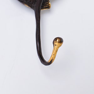 May include: A wall hook featuring a dark brown, textured metal base with a curved, elongated arm. The tip of the hook is finished in a polished, bright gold tone, creating a distinct contrast against the darker, matte-finished metal body.