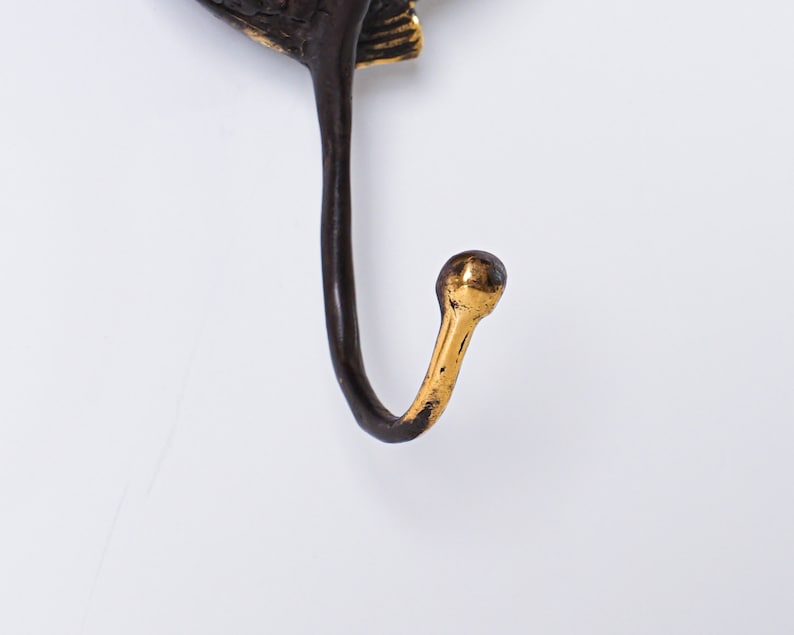 May include: A wall hook featuring a dark brown, textured metal base with a curved, elongated arm. The tip of the hook is finished in a polished, bright gold tone, creating a distinct contrast against the darker, matte-finished metal body.