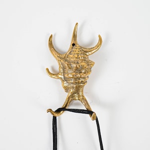 May include: A gold-coloured seashell-shaped wall hook with a black cord attached. The hook is designed to resemble a seashell with intricate details.