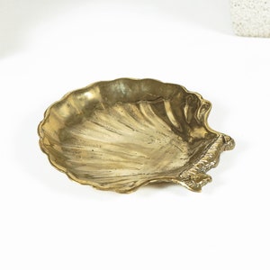 May include: A brass dish shaped like a scallop shell with a metallic gold finish. The dish features a textured, ribbed surface that mimics the natural ridges of a seashell, with a scalloped edge and a detailed hinge area at the side.