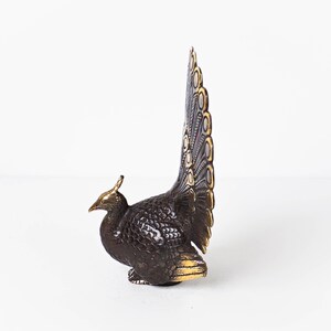 May include: A metallic peacock figurine featuring a dark bronze-toned body with textured feather detailing. The bird has a tall, upright tail fan with gold-toned accents on the edges and crest, showcasing a contrast between the dark and bright metal.