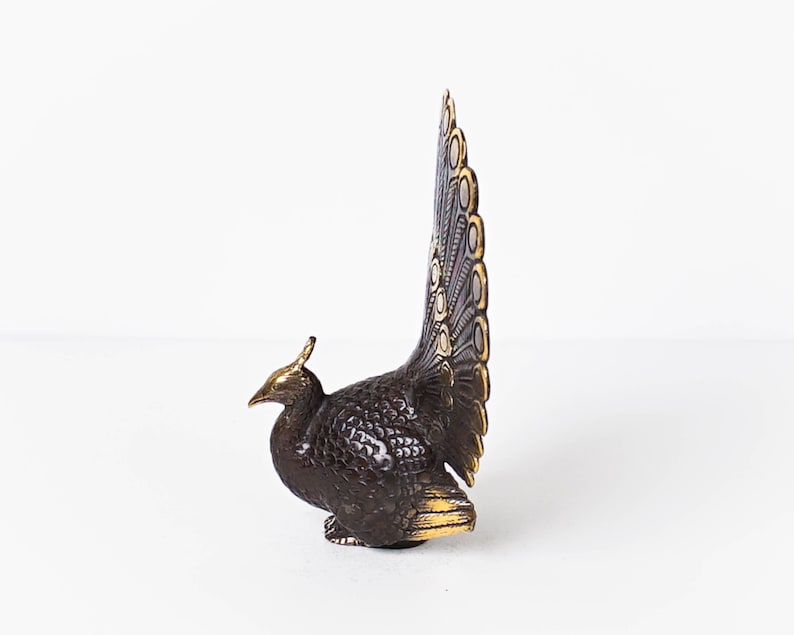 May include: A metallic peacock figurine featuring a dark bronze-toned body with textured feather detailing. The bird has a tall, upright tail fan with gold-toned accents on the edges and crest, showcasing a contrast between the dark and bright metal.