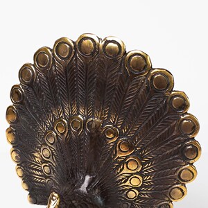 May include: A metallic bronze-toned peacock figurine featuring an expansive, semi-circular tail display. The fan-shaped tail is intricately detailed with embossed feather patterns and raised circular eye motifs arranged in a radial, symmetrical design.