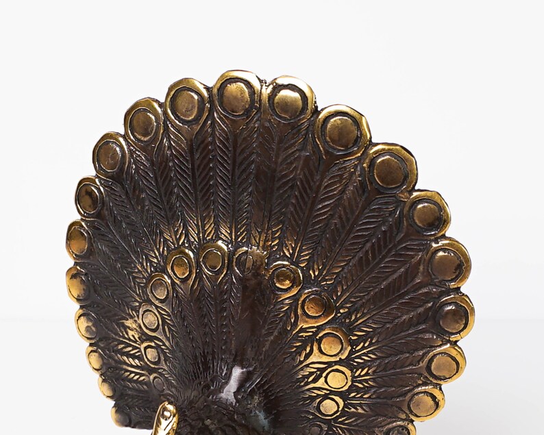 May include: A metallic bronze-toned peacock figurine featuring an expansive, semi-circular tail display. The fan-shaped tail is intricately detailed with embossed feather patterns and raised circular eye motifs arranged in a radial, symmetrical design.