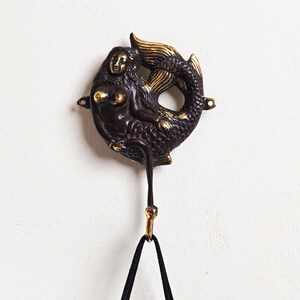 May include: A dark bronze-toned metal wall hook featuring a sculpted mermaid figure with polished gold-coloured accents on the hair, chest, and tail fin. The mermaid is positioned horizontally with a curved hook extending downward from the base.