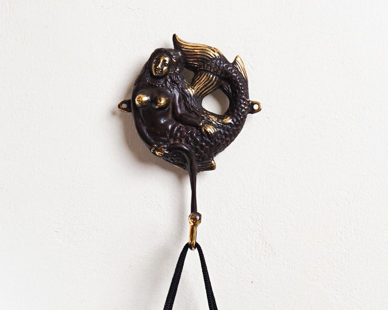 May include: A dark bronze-toned metal wall hook featuring a sculpted mermaid figure with polished gold-coloured accents on the hair, chest, and tail fin. The mermaid is positioned horizontally with a curved hook extending downward from the base.