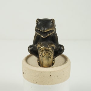 May include: A bronze frog figurine playing a drum. The frog is dark bronze with a gold-colored drum. The figurine sits on a round, off-white base. The drum has a geometric pattern.
