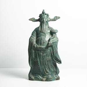 Peut inclure: Statue en bronze vieilli d'une figure vêtue d'une robe, avec une longue barbe et une coiffe élaborée. La statue tient un bol et présente des détails complexes sur les vêtements. La figure a une expression sereine et se tient debout.