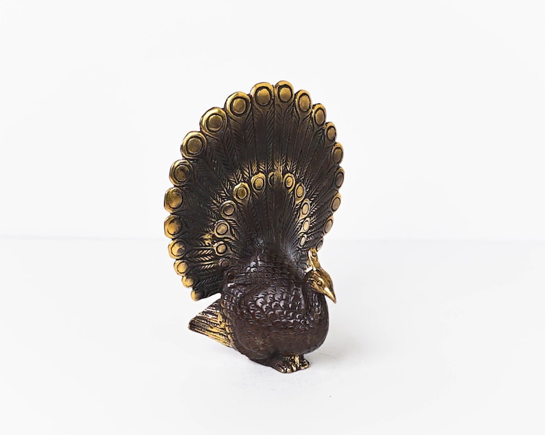 May include: A dark bronze-toned metal peacock figurine featuring a fanned tail with circular eye-like patterns. The bird has a textured body, a small head with a pointed beak, and a crest, with polished gold-colored highlights on the tail edges.