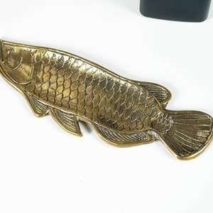 Peut inclure: Un plateau en m&eacute;tal dor&eacute; en forme de poisson, pr&eacute;sentant des &eacute;cailles, des nageoires et une queue en relief d&eacute;taill&eacute;es. La surface poss&egrave;de une finition textur&eacute;e et r&eacute;fl&eacute;chissante avec des lignes d&eacute;finies soulignant la t&ecirc;te et le corps.
