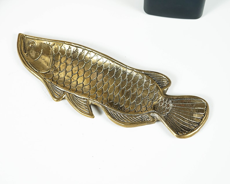 Peut inclure: Un plateau en m&eacute;tal dor&eacute; en forme de poisson, pr&eacute;sentant des &eacute;cailles, des nageoires et une queue en relief d&eacute;taill&eacute;es. La surface poss&egrave;de une finition textur&eacute;e et r&eacute;fl&eacute;chissante avec des lignes d&eacute;finies soulignant la t&ecirc;te et le corps.