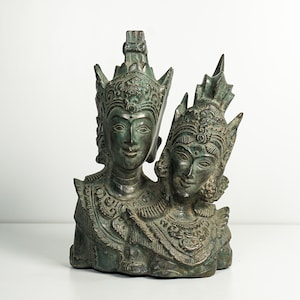 Peut inclure: Une sculpture en bronze aux tons vert foncé représentant deux personnages côte à côte. Chaque figure porte une coiffe pointue ornée et des bijoux de col détaillés. Les visages présentent des expressions calmes avec des yeux en amande.