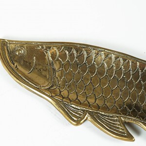 Peut inclure: Un plateau en laiton dor&eacute; en forme de poisson, pr&eacute;sentant des &eacute;cailles en relief d&eacute;taill&eacute;es, un &oelig;il d&eacute;fini et des nageoires textur&eacute;es le long du bord inf&eacute;rieur. La surface m&eacute;tallique a une finition polie avec des lignes sombres.
