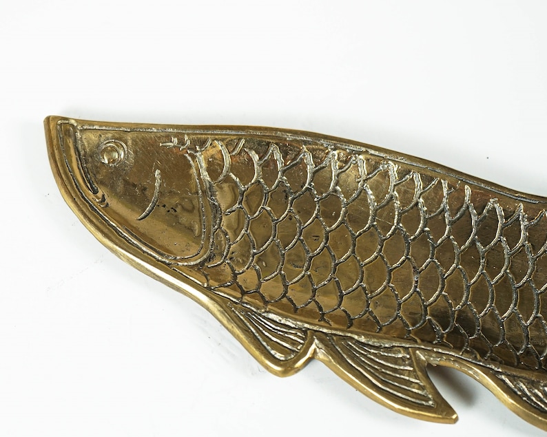 Peut inclure: Un plateau en laiton dor&eacute; en forme de poisson, pr&eacute;sentant des &eacute;cailles en relief d&eacute;taill&eacute;es, un &oelig;il d&eacute;fini et des nageoires textur&eacute;es le long du bord inf&eacute;rieur. La surface m&eacute;tallique a une finition polie avec des lignes sombres.