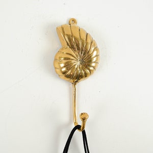 Könnte beinhalten: Ein goldfarbener Haken in Form einer Muschel, der an einer weißen Wand montiert ist.