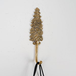 May include: A gold metal hook shaped like a pine tree. The hook is mounted on a white wall.