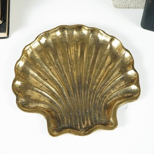 May include: A gold-toned brass dish shaped like a scallop shell. The dish features a textured, ribbed surface with a metallic finish and a scalloped edge, reflecting light across its curved, concave form. The object has a weathered patina.