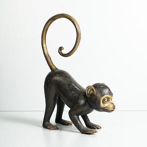 Puede incluir: Una figura de mono de color bronce oscuro de pie sobre cuatro patas con una cola larga y curvada que apunta hacia arriba. El mono presenta una cara, orejas y pies de tono dorado pulido más claro, con pelaje texturizado detallado.