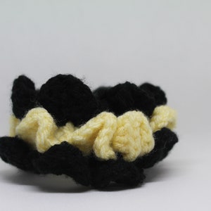 May include: A handmade crochet scrunchie in a flower shape. The scrunchie features alternating petals of black and cream-colored yarn. The accessory is designed to hold hair in a bun or ponytail.