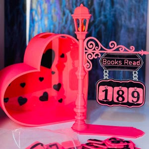 May include: A vibrant pink reading tracker set. The set includes a heart-shaped container with black heart cutouts, a street lamp-style sign that reads "Books Read" with the number 189, and small number tiles. The lamp post has a decorative design.