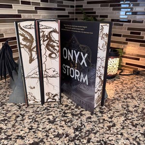 Op de afbeelding: Een zwart hardback boek getiteld "ONYX & STORM" van Rebecca Yarros, een best verkopende auteur. De boekomslag heeft een gouden draakontwerp en de tekst "Believe fate has carved some awesome dragons!" en "Bound beautiful half of unique magic!"
