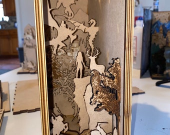 Peut inclure: Boîte d'ombre en bois représentant une scène fantastique avec des dragons, un château, des cerfs et des personnages à cheval. La boîte a une conception en couches, créant de la profondeur. Le cadre est de couleur marron clair, et la scène est finement découpée dans du bois.