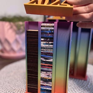 以下が含まれることがあります： 虹色のグラデーションデザインの3Dプリントブックエンド。ブックエンドには2つの棚があり、本が積み重ねられています。ブックエンドの上部には「2024」と書かれた取り外し可能な部分があります。