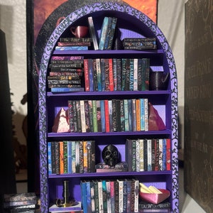 May include: A miniature purple bookshelf with five shelves, filled with a variety of fantasy and young adult novels. The bookshelf is decorated with a skull, a crystal, and a candlestick.