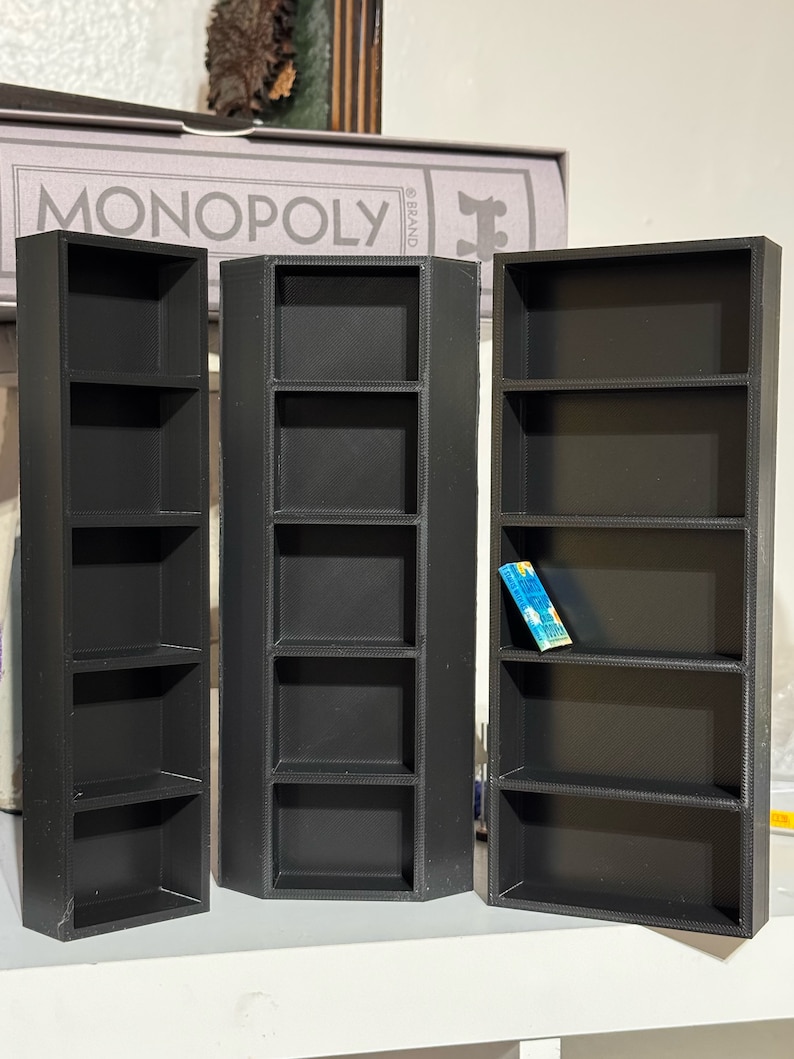May include: Three black, rectangular, open-shelf storage units with five shelves each. The units are made of a textured plastic material. The middle unit is slightly angled. A small blue and white card is on the top shelf of the rightmost unit.