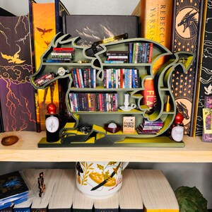 May include: A green dragon-shaped bookshelf with multiple shelves filled with books. The bookshelf is on a wooden shelf with a white mug with a floral design and a stack of books with the title "MAAS" on the spines.