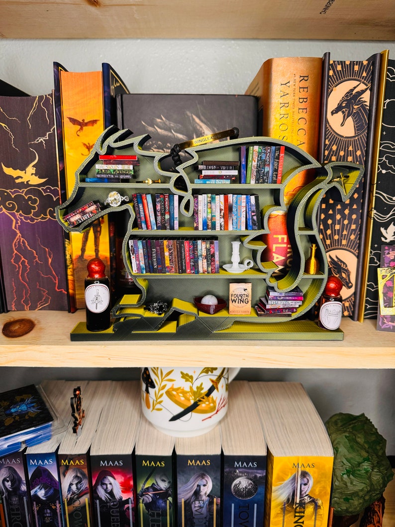 May include: A green dragon-shaped bookshelf with multiple shelves filled with books. The bookshelf is on a wooden shelf with a white mug with a floral design and a stack of books with the title "MAAS" on the spines.